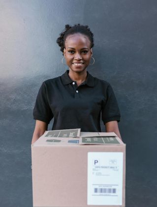A cheerful delivery woman in a black uniform holds a package outdoors.